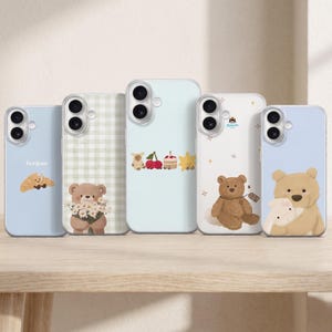 Könnte beinhalten: Fünf transparente Smartphone-Hüllen mit verschiedenen Illustrationen. Die Designs zeigen ein Croissant mit dem Text bonjour, einen Bären mit Gänseblümchen auf grünem Vichy-Karo, kleine Symbole von Bär, Kirschen, Kuchen und Stern, einen Bären mit Anhänger sowie einen Bären, der ein Kaninchen umarmt.