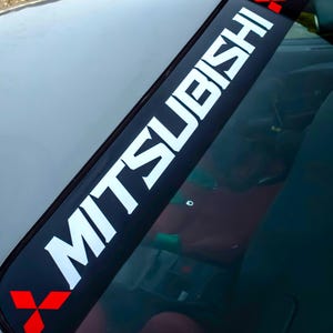 Może przedstawiać: Czarny baner na przednią szybę z białym napisem "MITSUBISHI" jest wyświetlany u góry przedniej szyby samochodu. Czerwone logo w kształcie rombu znajduje się na każdym końcu banera. Widoczna jest szara maska i wnętrze samochodu.