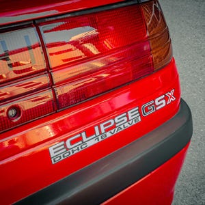 Peut inclure: Gros plan de l'arrière d'une voiture rouge, mettant en évidence le feu arrière et l'emblème "ECLIPSE GSX". Le texte "DOHC 16 VALVE" est également visible. La carrosserie de la voiture est rouge vif, avec un pare-chocs noir.