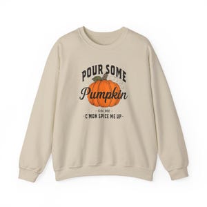 May include: A cream-colored sweatshirt with the text "Pour Some Pumpkin" above a graphic of an orange pumpkin. Below the pumpkin, the text reads "-ON ME- C'MON SPICE ME UP-". The sweatshirt has long sleeves and a crew neck.