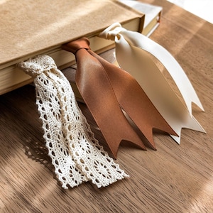 May include: Three bookmarks inserted into a closed tan book. One bookmark is made of white crocheted lace, one is a smooth copper-colored satin ribbon, and one is a smooth cream-colored satin ribbon. Each ribbon bookmark has a V-shaped cut end.
