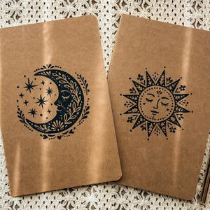 May include: Two rectangular notebooks with tan covers. The left notebook features a black ink illustration of a crescent moon with a face, stars, and floral details. The right notebook displays a black sun illustration with a face and rays.