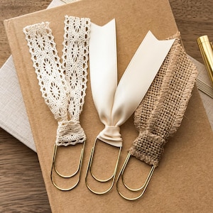 May include: Three large gold-toned metal paper clips, each adorned with a different fabric topper. One features cream-colored intricate lace, the second has a smooth off-white satin ribbon, and the third is crafted from a textured brown burlap fabric.