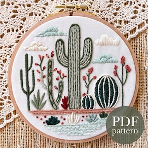 Può includere: Arte di ricamo a cerchio che rappresenta un paesaggio desertico con cactus, piante grasse e nuvole. Il cerchio è in legno e ha uno sfondo in tessuto bianco. Il ricamo è realizzato in tonalità di verde, rosso e marrone.