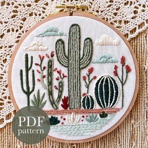 May include: Embroidered cactus scene in a wooden hoop. The design features various cacti, red flowers, and blue and white clouds. The text "PDF pattern" is visible on the bottom left. The embroidery is on a white fabric.
