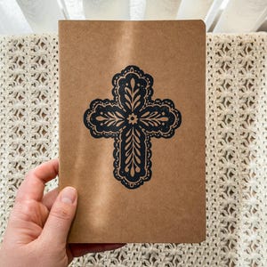 May include: A rectangular notebook with a light brown kraft paper cover featuring a centered black ink stamp of an ornate cross. The cross design includes intricate filigree-style leaf patterns and a scalloped border, creating a bold contrast.