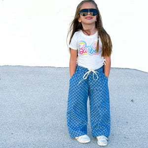 Può includere: Una persona giovane indossa occhiali da sole, una maglietta bianca con la scritta "Barbie" in caratteri colorati e pantaloni blu a scacchi. I pantaloni hanno una vita con coulisse e gambe larghe. Le scarpe da ginnastica bianche completano l'outfit.