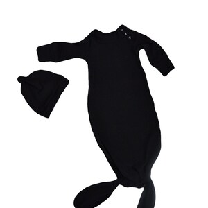 Puede incluir: Un pelele negro para bebé con mangas largas y un gorro a juego. El pelele tiene la parte inferior anudada y el gorro tiene la parte superior puntiaguda.