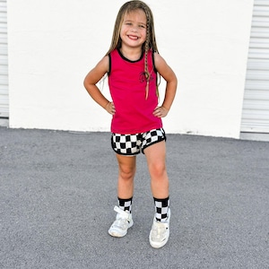 May include: A young person wearing a magenta tank top with black trim and a small graphic, paired with black and white checkered shorts. The outfit is completed with white sneakers and black and white checkered socks. The person is smiling.