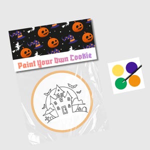 Puede incluir: Un kit de decoración de galletas con temática de Halloween con una galleta redonda de azúcar, una bolsa de plástico y una paleta de pintura con cuatro colores. La galleta tiene un contorno en blanco y negro de una casa encantada con luna llena y murciélagos. La paleta de pintura tiene amarillo, verde, morado y naranja. La bolsa tiene una etiqueta que dice "Pinta tu propia galleta" con un patrón repetido de calabazas naranjas y sombreros de bruja.