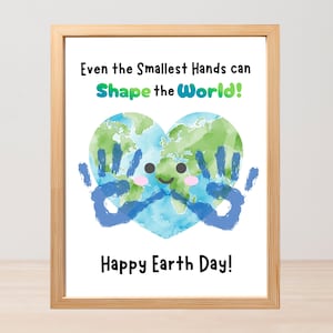 May include: Framed print with a light wood grain frame. The artwork features a watercolor illustration of Earth in a heart shape, with blue handprints and a smiling face. Text reads "Even the Smallest Hands can Shape the World!" and "Happy Earth Day!"