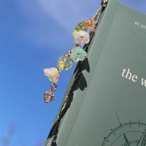 May include: A gold-colored metal bookmark with a chain of colorful beads and flower charms. The bookmark is attached to a book with the words "the way" visible. The background is a bright blue sky.