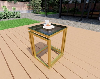 May include: A square side table featuring a gold-colored metal frame with a geometric base and a dark glass top. A white ceramic cup and a small white plate with a pastry are placed on the surface of the table, which stands on a wooden deck.