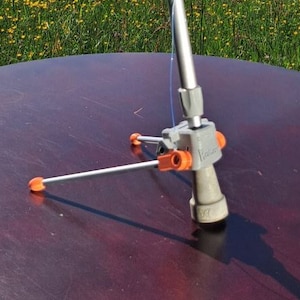 Könnte beinhalten: Eine graue Unterarmstütze mit einem orangefarbenen verstellbaren Griff und einem Dreibeinstativ. Die Krücke steht auf einem dunkelbraunen Tisch mit einem grünen Feld und Bergen im Hintergrund.