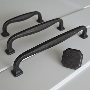 Puede incluir: Tres tiradores de armario de metal negro con acabado desgastado. Los tiradores son de diferentes tamaños y están dispuestos en fila sobre una superficie blanca. También se muestra un pomo de metal negro a juego.