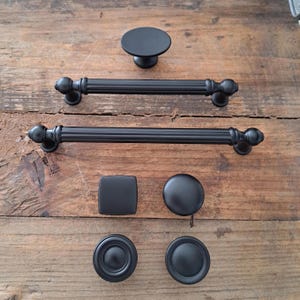 May include: A set of black cabinet hardware, including two drawer pulls, four knobs, and one larger knob. The knobs are round, square, and have a decorative design. The drawer pulls are long and have a simple design.