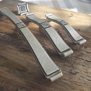 May include: Three silver-toned metal cabinet pulls of varying lengths and one square cabinet knob. Each piece features a matte finish with a central decorative panel displaying a raised, cross-hatched geometric pattern in a darker silver tone.