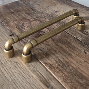 Może przedstawiać: Dwa uchwyty do szafek ze starego mosiądzu o konstrukcji przypominającej rury. Każdy uchwyt ma prosty pręt połączony z zakrzywionymi końcami i cylindrycznymi podstawami. Okucia mają ciepły, postarzany mosiężny kolor, sugerujący styl vintage.