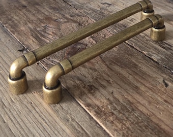 May include: Two antique brass cabinet pulls with a pipe-like design. Each pull has a straight bar connected to curved ends and cylindrical bases. The hardware is a warm, aged brass color, suggesting a vintage aesthetic.