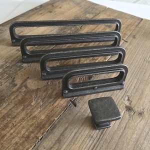 May include: Four dark gray metal drawer pulls and one square knob are arranged on a rustic wooden surface. The rectangular pulls have a curved handle design. The knob is square with a short, rectangular base. Hardware for cabinets or furniture.