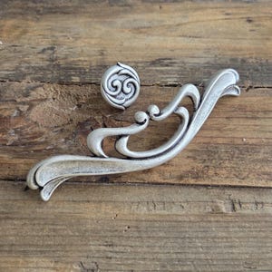 Pode incluir: Conjunto de ferragens decorativas prateadas, incluindo um puxador e uma pega curva. O puxador apresenta um design em espiral, enquanto a pega tem uma forma ornamentada e fluida. As ferragens são exibidas numa superfície de madeira rústica.