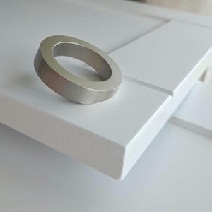 Może przedstawiać: Uchwyt pierścieniowy ze szczotkowanego niklu spoczywa na białej powierzchni. Okrągły uchwyt ma nowoczesny, minimalistyczny design. Biała powierzchnia wydaje się być częścią frontu szafki lub szuflady.
