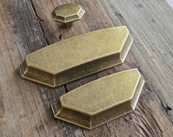 May include: A set of antique brass cabinet hardware, including two rectangular pulls and a hexagonal knob. The hardware has a weathered finish, suggesting an aged aesthetic. The pieces are arranged on a rustic wooden surface.