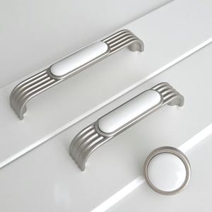 May include: Three white ceramic cabinet knobs with a silver metal base. The knobs have a rounded rectangular shape and a grooved design.