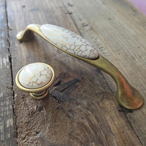 Op de afbeelding: Een set meubelbeslag, bestaande uit een handgreep en een knop. De handgreep en knop hebben een ovale, witte kern met een gouden craquelé-patroon. Deze decoratieve elementen zijn geschikt voor meubels.