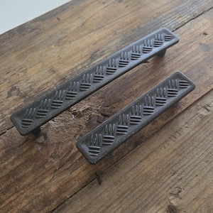 May include: Two black metal cabinet pulls with a geometric pattern. The pulls are rectangular and have a slightly curved shape.