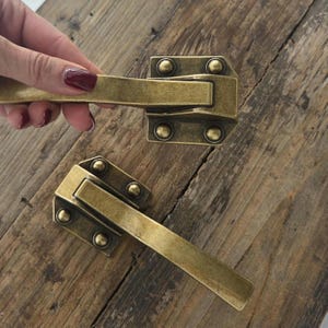 May include: Two antique brass-toned cabinet pulls with a rectangular design. Each pull has a handle and a rectangular base with four rivets. One pull is being held by a hand with dark red nail polish. The pulls are on a wooden surface.