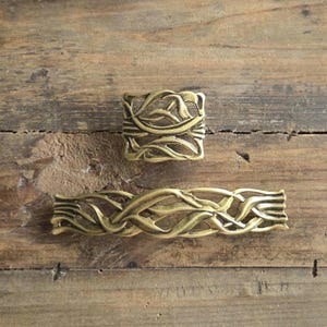 May include: Two antique brass cabinet pulls with a flowing leaf design. One is square, the other rectangular. The hardware is set against a rustic wooden background, showcasing the warm tones of the metal.