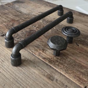 Peut inclure: Deux poignées de meubles en bronze foncé et deux boutons assortis reposent sur une surface en bois vieilli. Les poignées sont en forme de barre avec des extrémités incurvées. Un bouton est un disque plat avec des cercles concentriques, l'autre est hexagonal.
