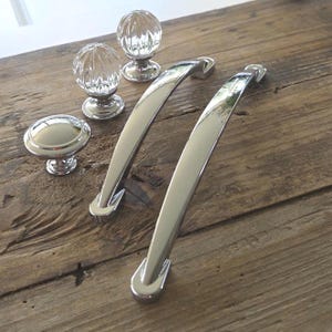 May include: A collection of cabinet hardware including two curved silver metal drawer pulls, one smooth round silver metal knob, and two clear glass spherical knobs with a ribbed texture, all featuring a polished and reflective metallic finish.