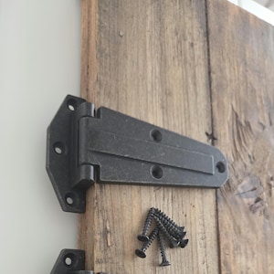 May include: Close-up of a black metal hinge attached to a weathered wooden surface. The hinge has multiple screw holes and is accompanied by a small pile of black screws. The wood has a natural, aged appearance.