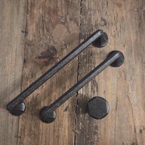 May include: Two black metal cabinet pulls and a matching knob rest on a weathered wooden surface. The pulls are rectangular with rounded ends, and the knob is a hexagon shape. The hardware has a rustic, industrial aesthetic.