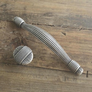 Puede incluir: Un tirador de armario y un pomo a juego de color plateado con un diseño acanalado. El tirador curvo mide aproximadamente 18 cm de largo y el pomo es redondo. Ambos están sobre una superficie de madera rústica.