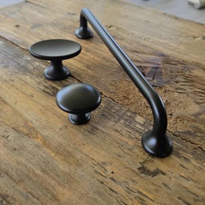 May include: A set of matte black cabinet hardware consisting of one long arched pull handle and two round knobs. The hardware features a smooth, solid finish with a curved design for the handle and a circular, pedestal-style base for the knobs.