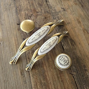 May include: Two elongated drawer pulls and two round knobs made of antique brass-toned metal. The pulls feature an oval white ceramic center with the text Vintage 1808 in black. One knob is plain brass, while the other has a white ceramic inlay.