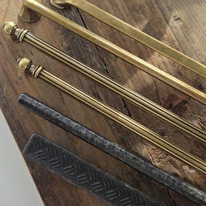 Puede incluir: Una selección de tiradores de metal para armarios en varios estilos y acabados. Los tiradores incluyen opciones de latón y negro, con diseños que van desde barras simples hasta patrones detallados, ranurados y texturizados. El hardware se muestra sobre una superficie de madera rústica.