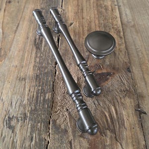 May include: Two dark bronze cabinet pulls and a matching knob rest on a weathered wooden surface. The pulls are long and slender with decorative detailing. The knob is round and flat. These hardware pieces are designed for furniture.