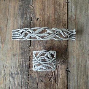 May include: Two decorative metal drawer pulls with a distressed, off-white finish. One is rectangular, the other square, both featuring an interwoven, organic design. The pulls are set against a rustic, weathered wooden surface.