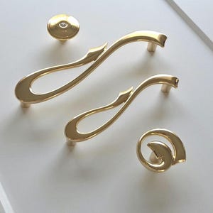 Op de afbeelding: Goudkleurige kastbeslagset. Bevat twee gebogen handgrepen, twee knoppen en een ronde trekker. De hardware heeft een glanzende afwerking en een decoratief, vloeiend ontwerp. De set wordt gepresenteerd op een wit oppervlak.