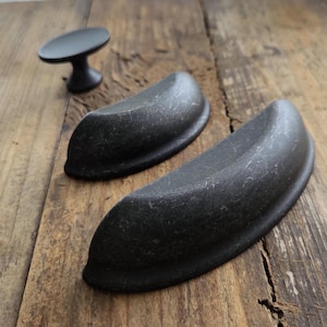 以下が含まれることがあります： 黒いキャビネット金具3点セット。2つはカッププルで、1つは小さなノブです。 金具は使い古したような仕上がりで、木製の表面に置かれています。