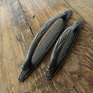 May include: Two dark metallic cabinet pulls with a weathered finish. Each handle features a central arched grip with a diamond-shaped embossed detail, mounted on a rectangular backplate with stepped, geometric ends and a distressed texture.
