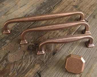 May include: Three copper-colored cabinet pulls and a matching knob rest on a weathered wooden surface. The pulls have a curved handle design with square bases. The knob is an octagon shape. The hardware has a rustic, antique appearance.