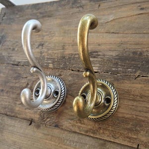 May include: Two wall-mounted hooks with a curved, S-shaped design and a circular base featuring a rope-like border. One hook has a matte silver finish, while the other has an antique brass finish. Each base includes two visible screw holes for mounting.