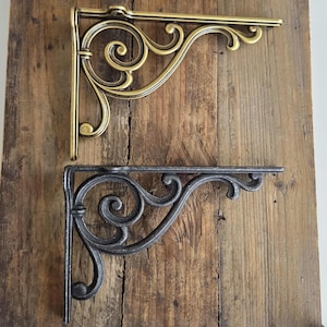 Puede incluir: Dos escuadras de estantería ornamentadas. Una es de color latón, la otra es gris oscuro. Ambas tienen un diseño de volutas decorativas. Los soportes están montados sobre una superficie de madera envejecida. Ideales para la decoración del hogar.