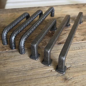 Könnte beinhalten: Eine Sammlung von sechs dunkelgrauen Metall-Schrankgriffen. Drei Griffe haben ein strukturiertes, rebar-ähnliches Design, während die anderen drei eine glatte, rechteckige Form haben. Die Griffe sind auf einer Holzoberfläche angeordnet.