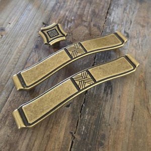 May include: A set of antique brass cabinet hardware, including two long pulls and a small knob. The hardware features a rectangular design with a geometric cross pattern, perfect for adding a vintage touch to furniture or cabinetry.
